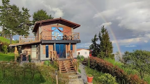 Ein zweistöckiges Haus mit Balkon, umgeben von üppigem Grün und einem Regenbogen im Hintergrund.