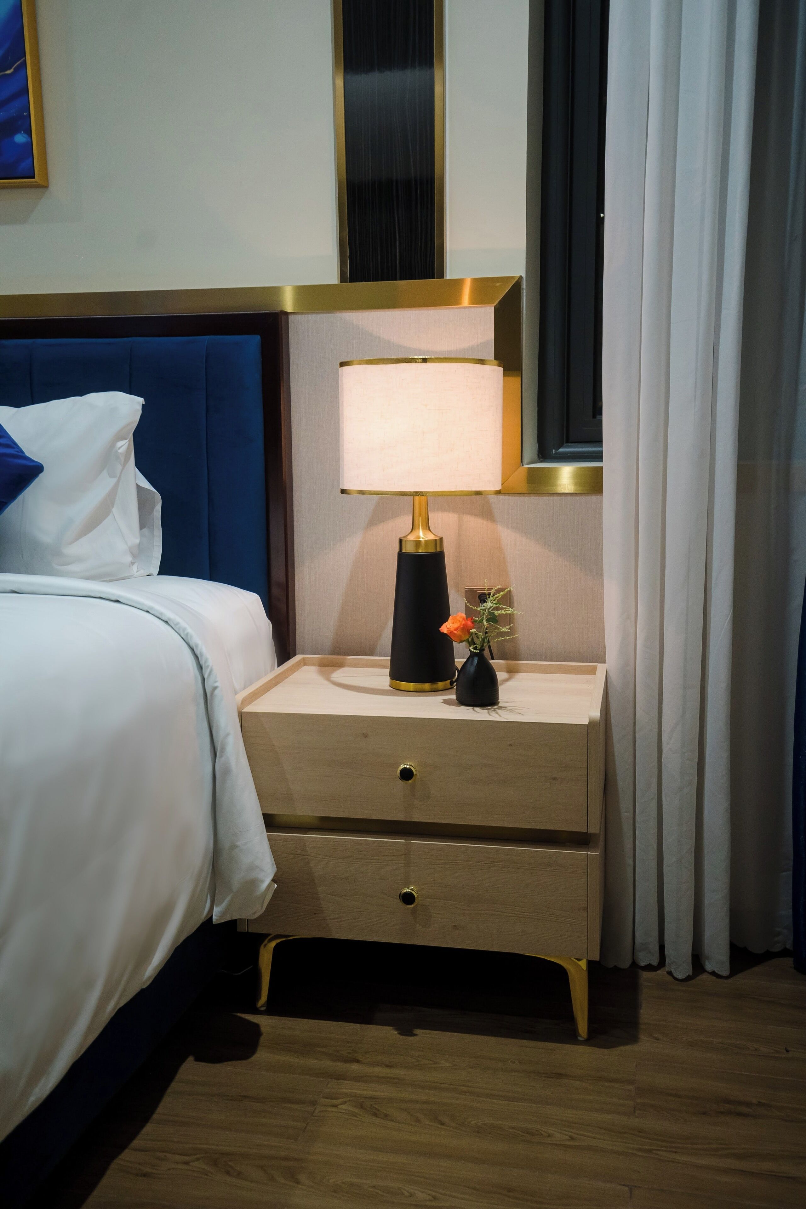 A neatly made bed with white linens and blue pillows, a bedside table with a lamp, a small vase with flowers, and a wooden nightstand.