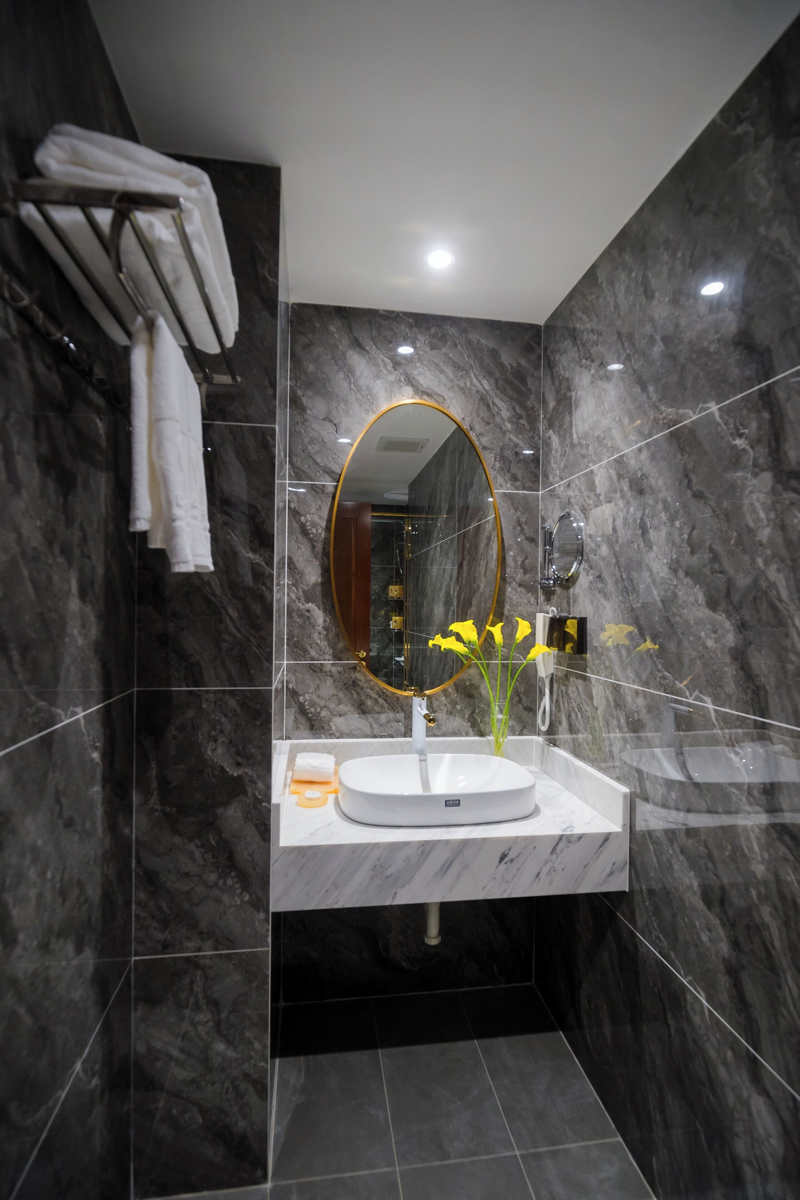 Une salle de bain moderne avec un mur en marbre, un miroir rond, un lavabo blanc et un vase rempli de fleurs jaunes.