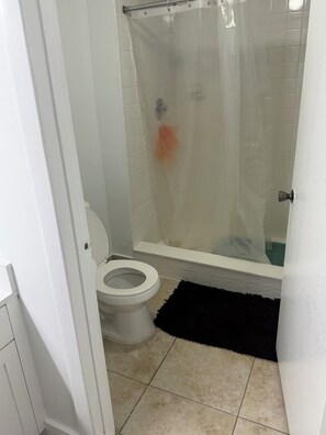 A bathroom with a toilet, a shower with a transparent curtain, and a black rug on the floor. - This is a two bed and two bath. Spacious Master Suite Near Hard Rock & Sawgrass. (Plantation)