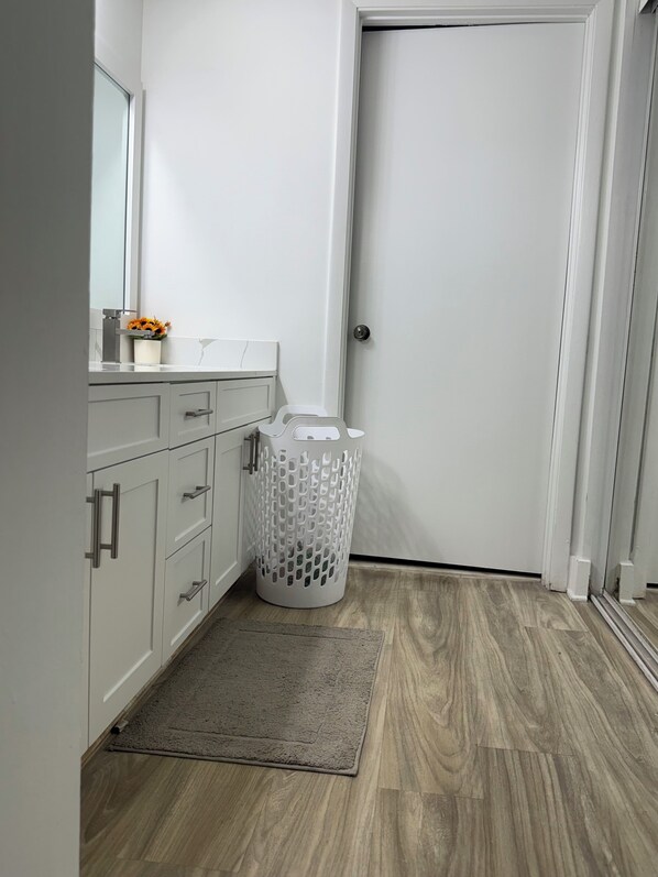 Un baño con lavamanos blanco, espejo y un pequeño arreglo floral. Se observa una cesta de ropa blanca y una puerta con manija plateada.