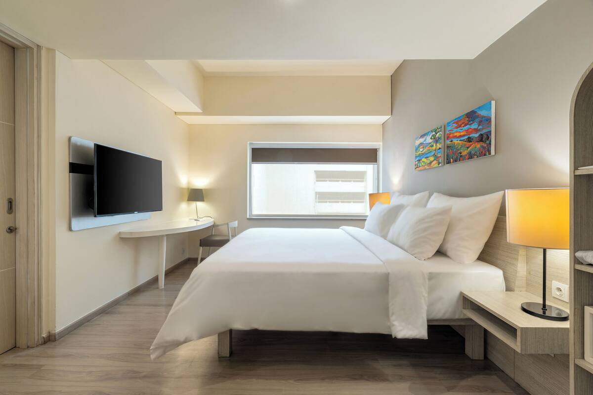 A modern hotel room with a large bed, a flat-screen TV mounted on the wall, a bedside table with a lamp, and a window with blinds.