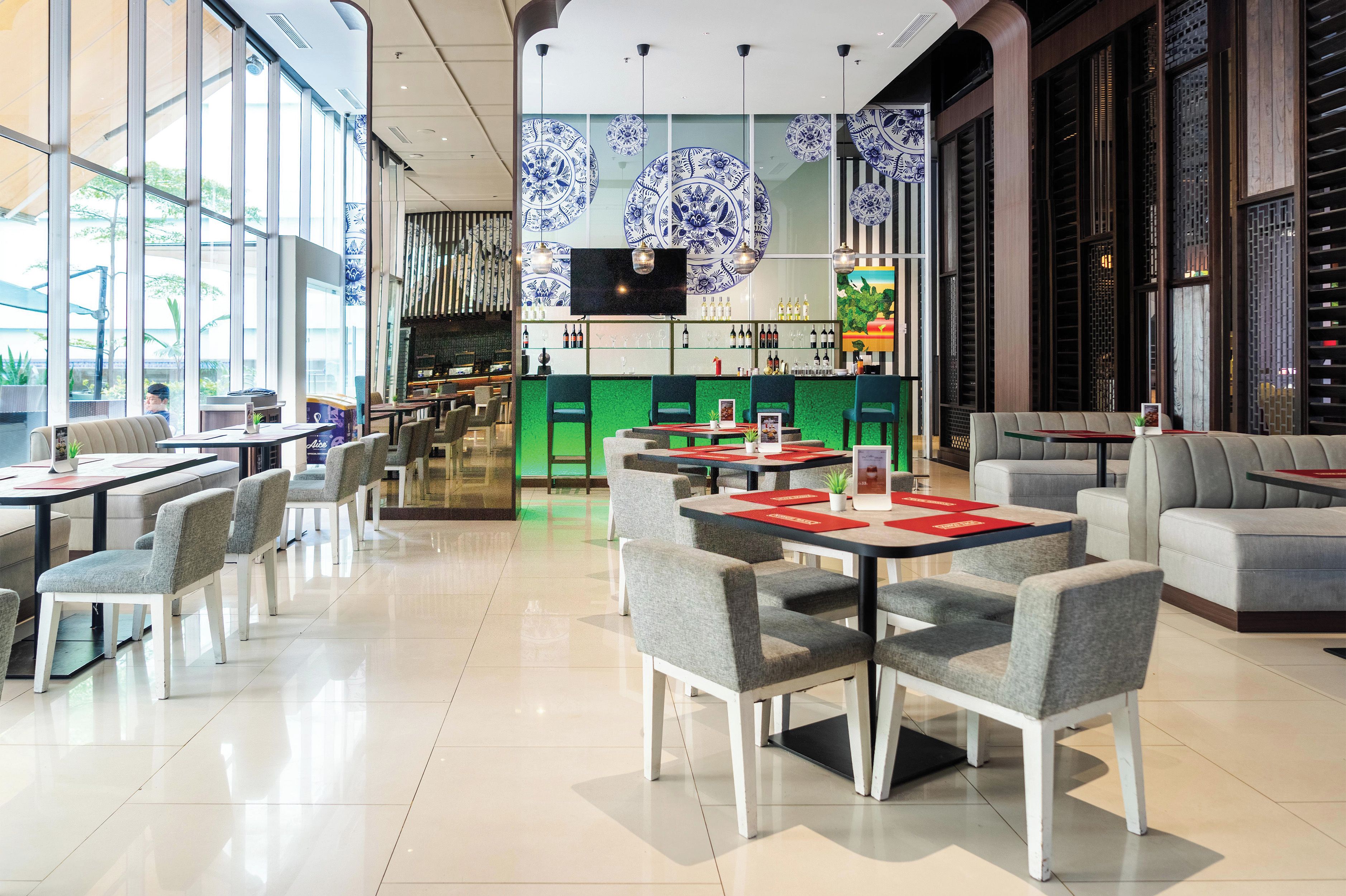 Interior moderno de restaurante con varias mesas y sillas, una zona de bar y grandes ventanales que dejan entrar luz natural.