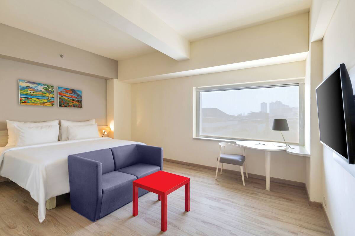A hotel room with a bed, a gray sofa, a red table, a TV, and two paintings on the wall.