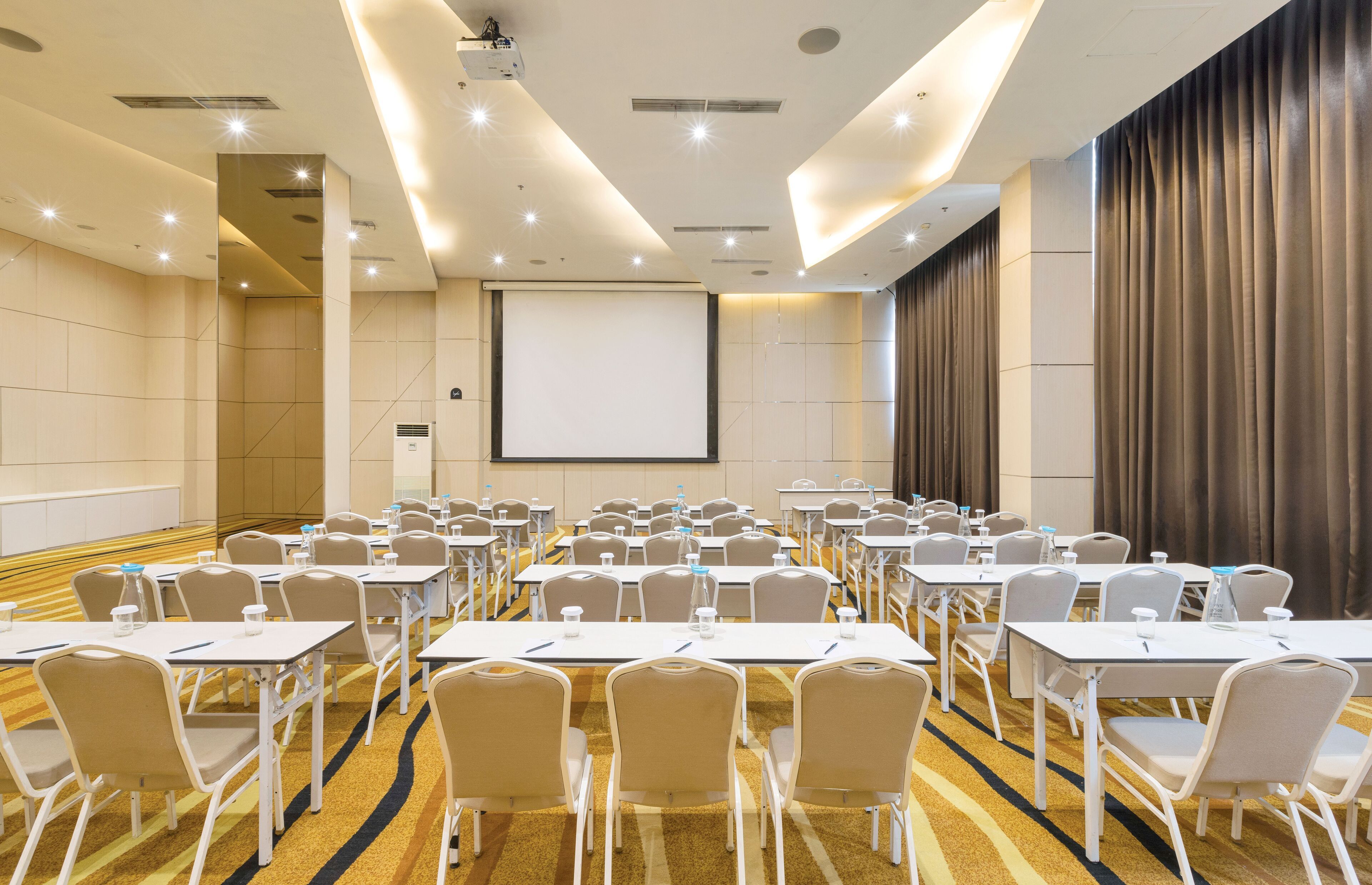 A conference room with rows of tables and chairs, a projector screen, and a large curtain.