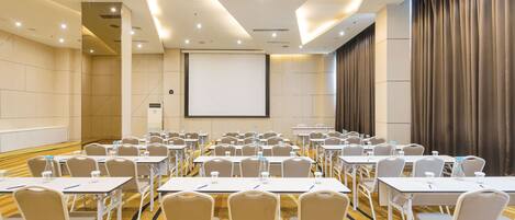 A conference room with rows of tables and chairs, a projector screen, and a large curtain.