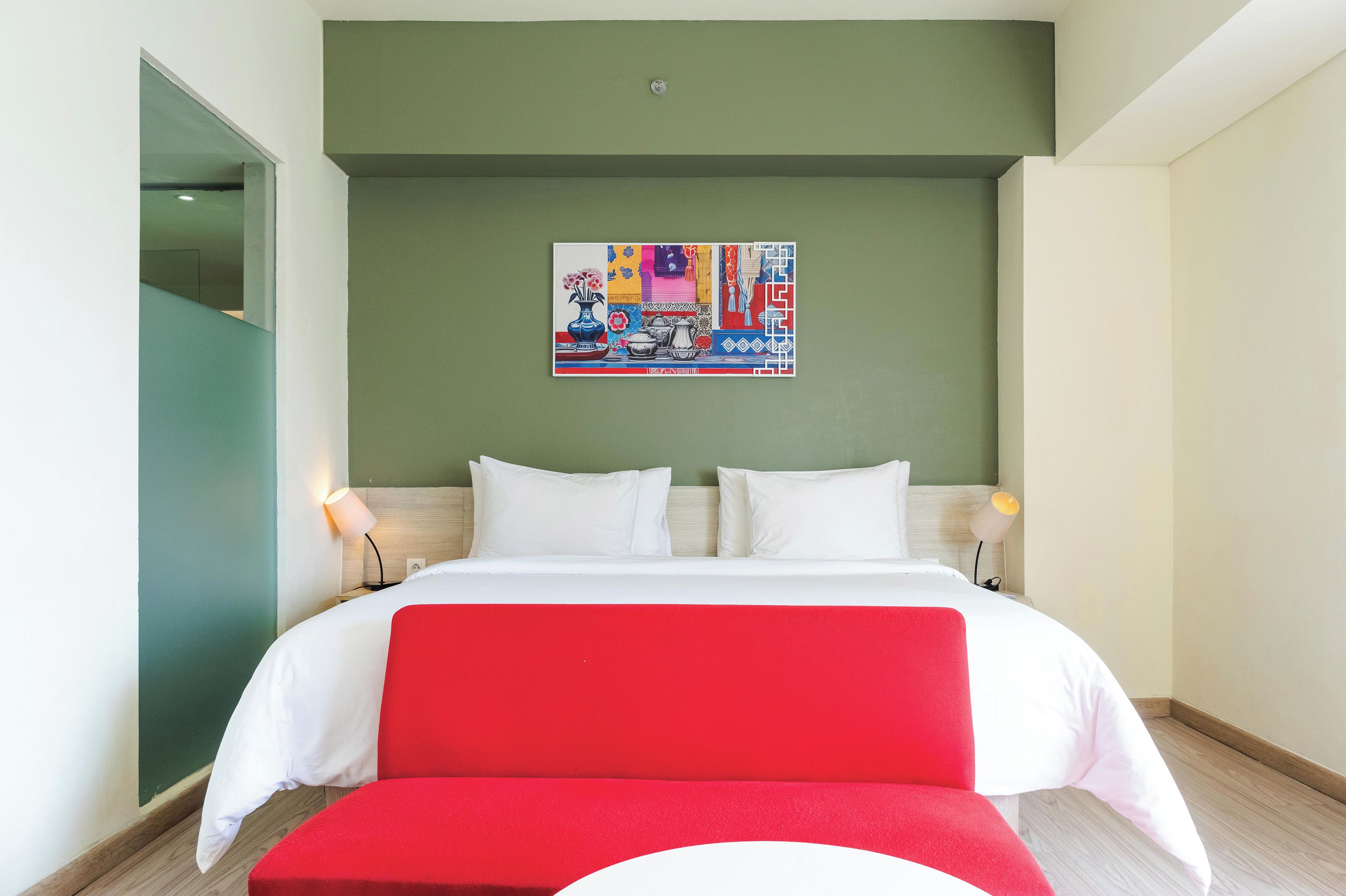 A hotel room with a bed, two bedside tables, a red bench, and a colorful artwork on the wall.