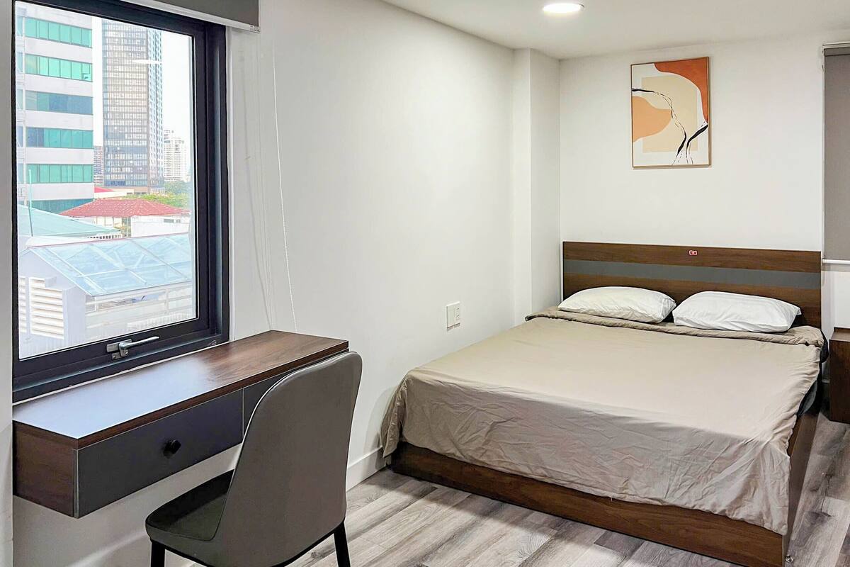 Ein Schlafzimmer mit einem Bett, einem Schreibtisch und einem Stuhl. Ein Fenster bietet Blick auf die Gebäude draußen.