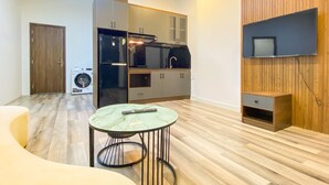 A compact living space with a kitchenette, a washing machine, a round table with a remote control, and a TV mounted on the wall. - Zeus Living - 7C Downtown By Feet (Ho Chi Minh City)