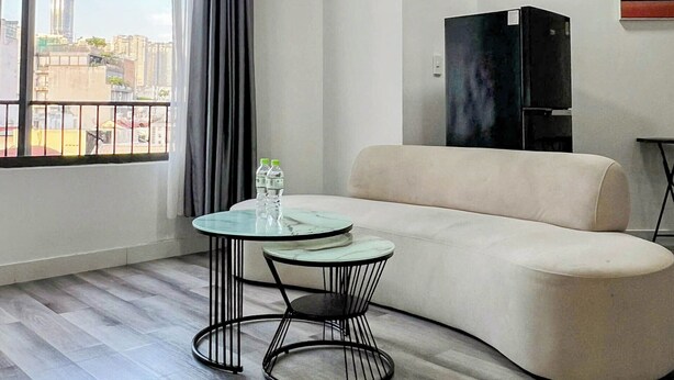 A modern living room with a curved sofa, a round glass table, and a view of a cityscape through large windows. - Zeus Living - 7C Downtown By Feet (Ho Chi Minh City)