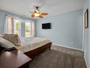 A bedroom with a bed, a ceiling fan, a TV mounted on the wall, and a window with curtains.