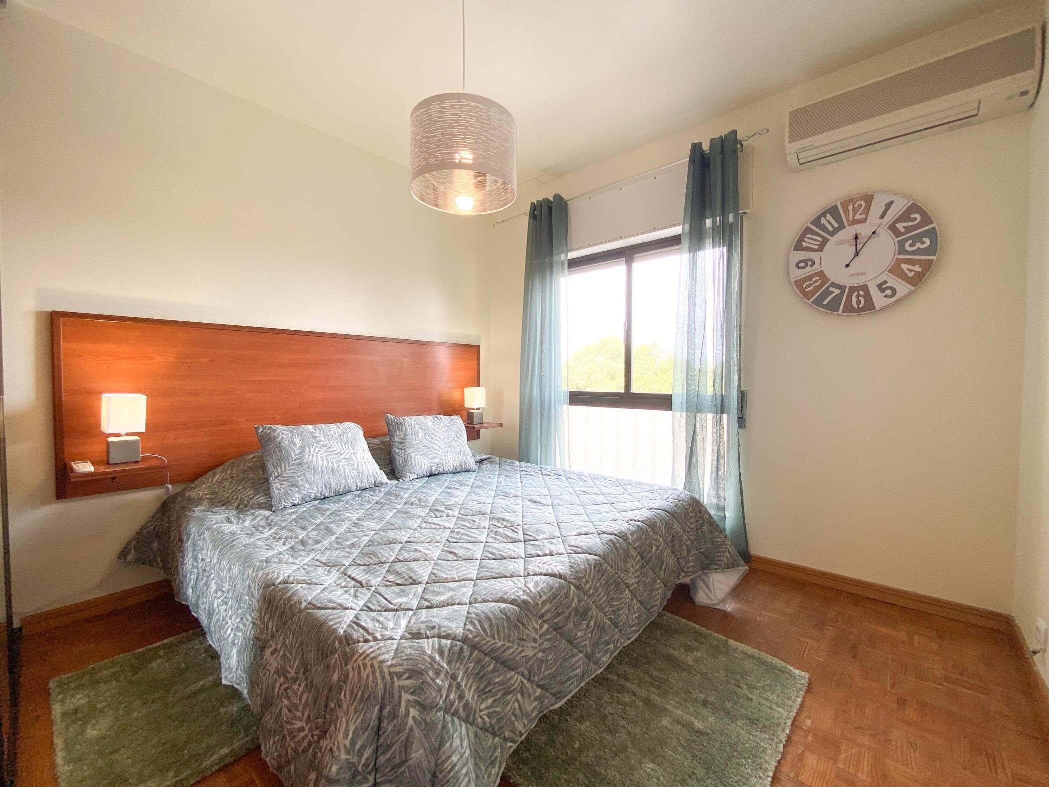 Une chambre avec une tête de lit en bois, un lit recouvert d’une literie à motifs, un tapis vert, une horloge murale et une fenêtre avec des rideaux.