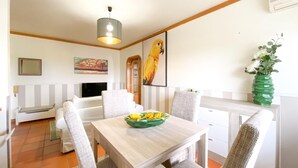 Zona pranzo con tavolo di legno, quattro sedie, una ciotola di frutta verde e un quadro di un pappagallo, più un vaso di fiori verdi.