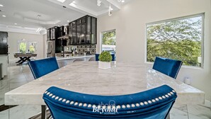 A modern kitchen with dark cabinetry, a central island, and a dining area with a marble table and blue upholstered chairs. - Sprawling Home w/ Rooftop Spot (San Antonio)