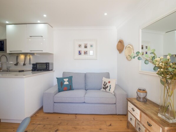 Un espacio habitable compacto con kitchenette, un sofá gris con cojines decorativos, una mesita de madera con un florero y un espejo en la pared.