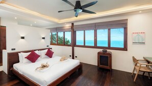 A hotel room with a large bed, wooden floor, a ceiling fan, a small table with chairs, and a window with a view of the sea. - Koh Tao Villas (Koh Tao)