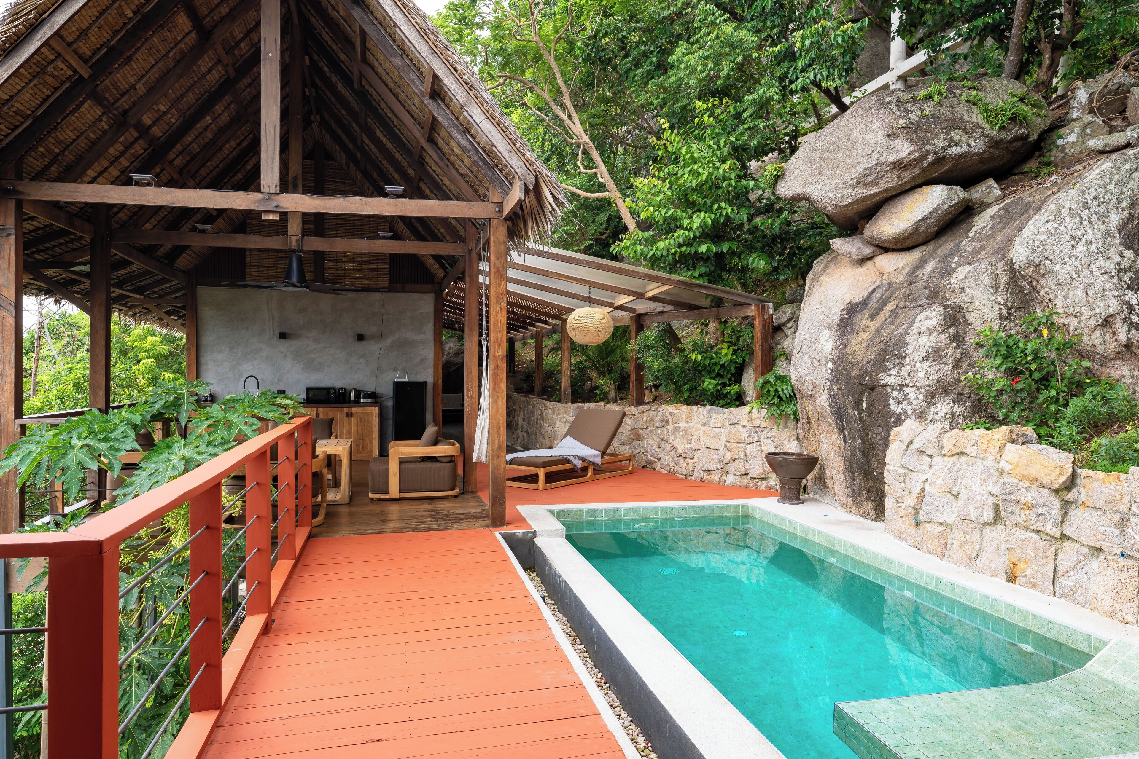 Um deck de madeira com piscina, cadeiras de descanso e uma parede de pedra.