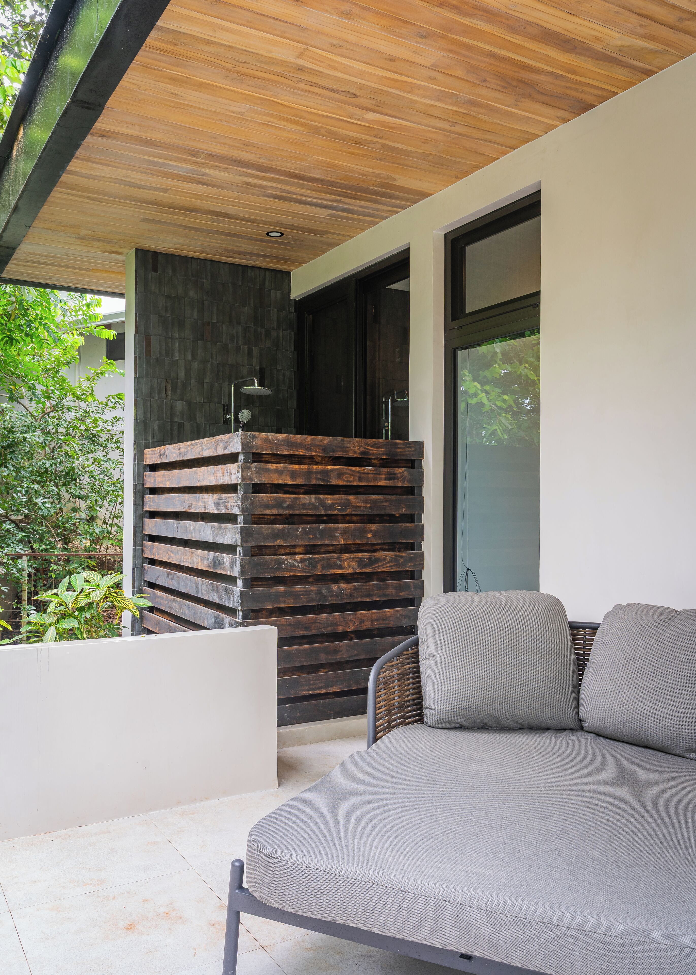 Un espace de détente extérieur moderne, doté d’un banc intégré et d’un canapé, d’un plafond en bois et d’une porte vitrée donnant sur un intérieur sombre.