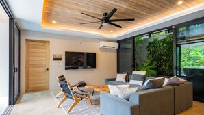 A modern living room with a grey sofa, a wooden ceiling fan, and a large TV. - Casa Pipeline: Jungle Villa, Pool, Sleeps 14 (Nosara)