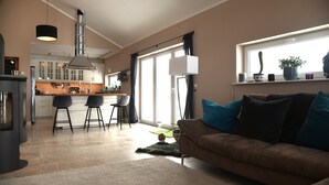Cozinha moderna com armários brancos, área de jantar com banquetas e sala de estar com sofá marrom e almofadas azuis.