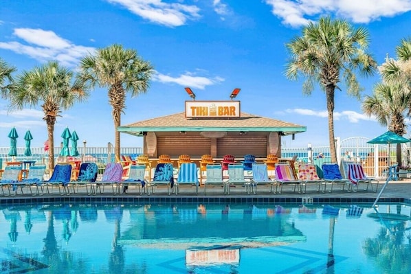 Un espace piscine doté d’un bar de style tiki, de chaises longues et de palmiers.