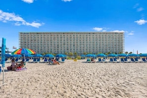 Une plage bordée de rangées de parasols bleus et de chaises longues, un grand bâtiment d’hôtel en arrière-plan et un ciel bleu dégagé.