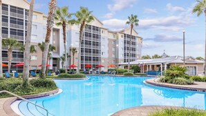 A swimming pool surrounded by palm trees and a hotel building with multiple floors and balconies.