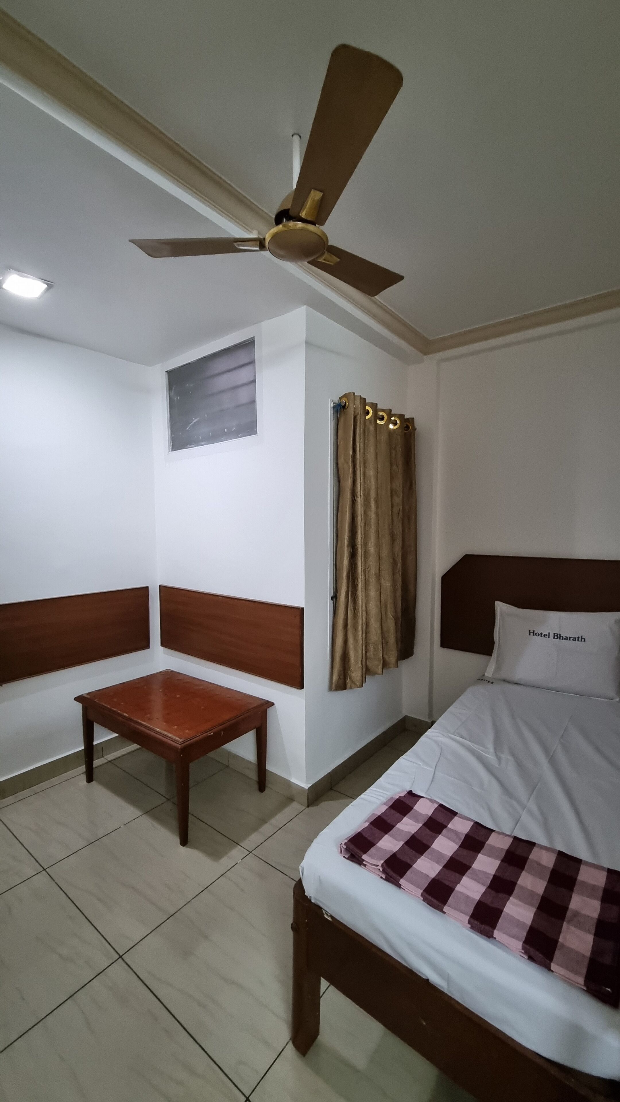A hotel room with a bed, a wooden nightstand, a ceiling fan, a window with curtains, and a wall-mounted light fixture.