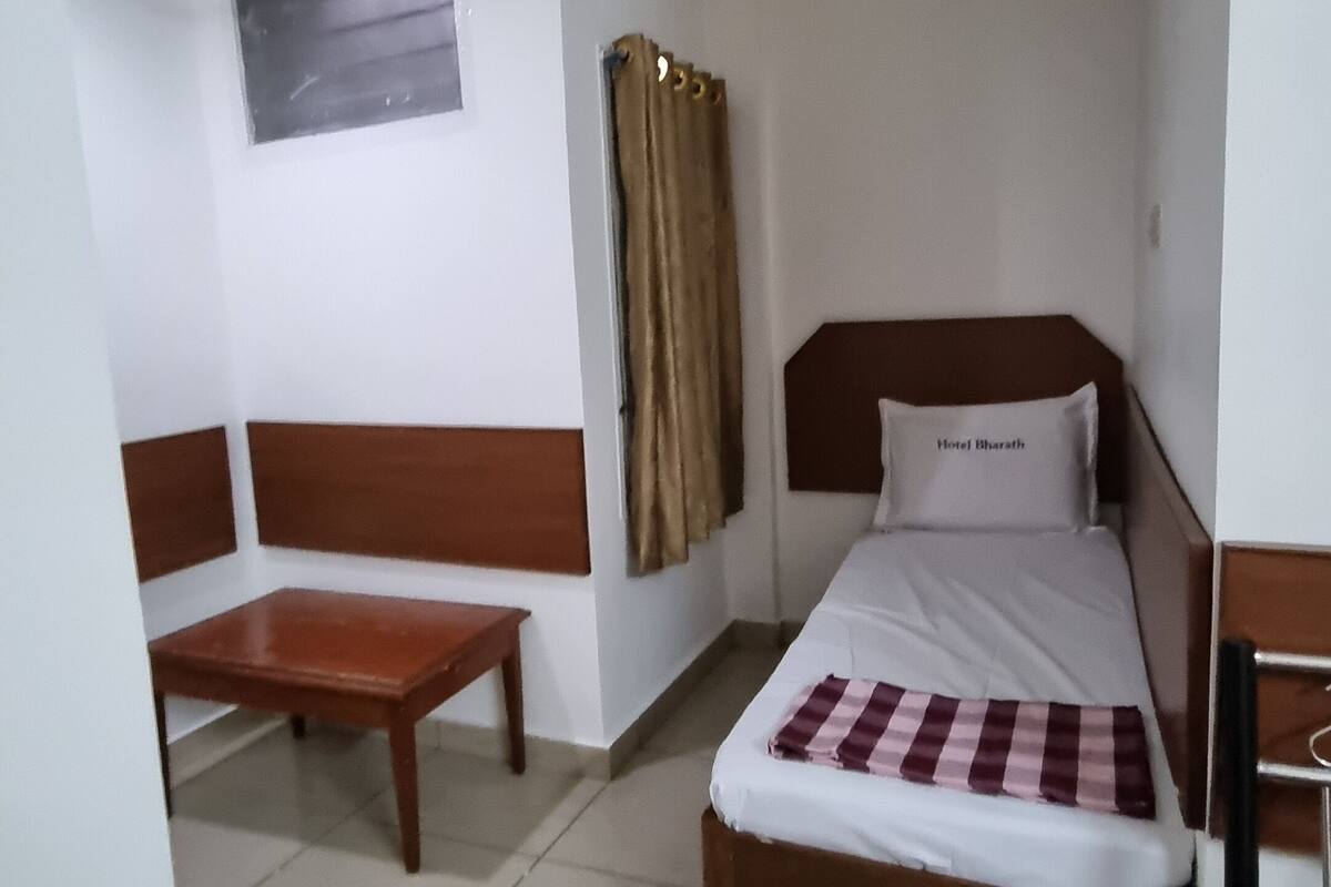 A single bed with a headboard, a wooden bench, a ceiling fan, a window with curtains, and a wall-mounted light switch.