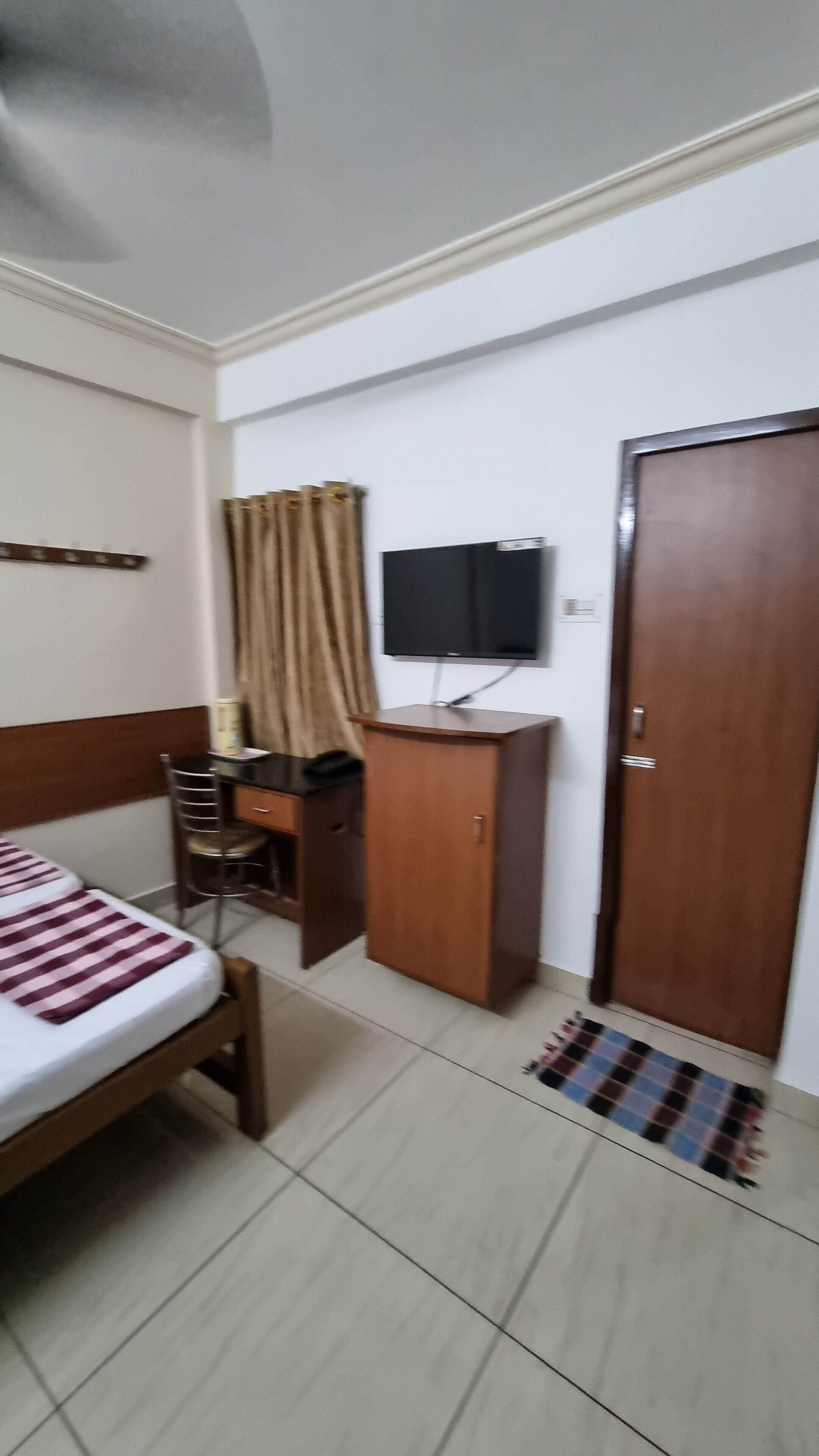 A hotel room with a bed, a desk with a chair, a television mounted on the wall, and a door.