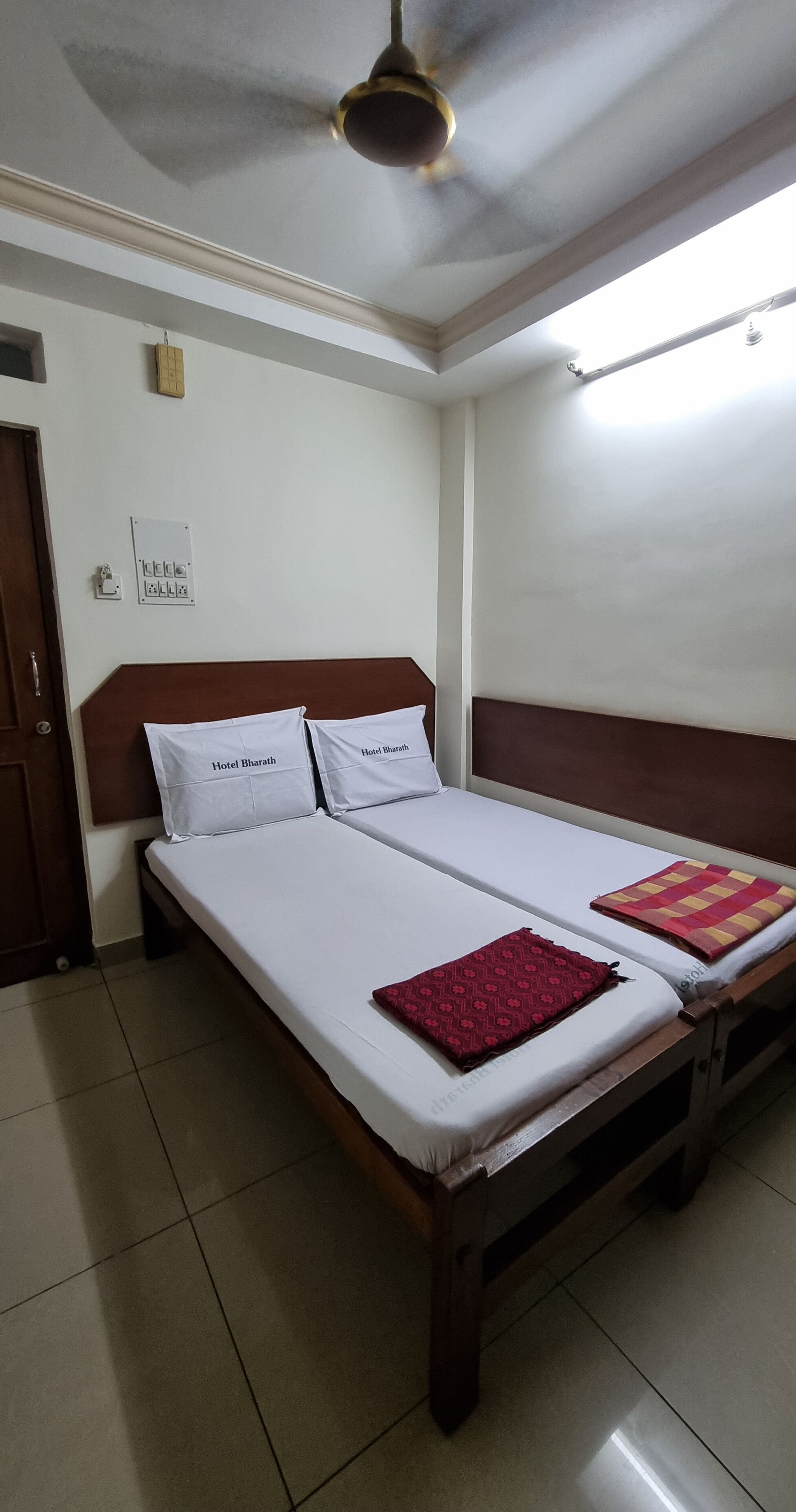 A double bed with white linens and two pillows, a wooden headboard, a red patterned throw, and a ceiling fan.