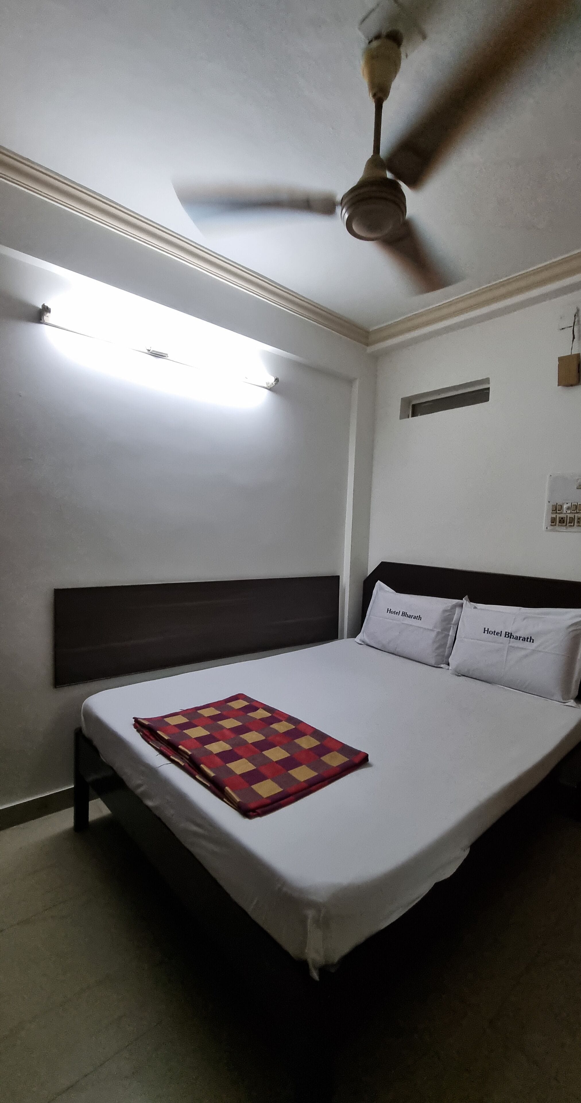 Ein Einzelbett mit Kopfteil, zwei Kissen mit dem Aufdruck 'Hotel Bharath', ein Deckenventilator und eine Leuchte über dem Bett.