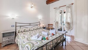 A bedroom with a bed, bedside tables, a nightstand, a chair, a window with curtains, and a wooden ceiling. - Casa Rolè (Dormelletto)