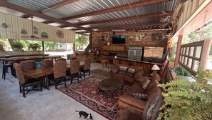 A rustic outdoor dining area with wooden furniture, a large rug, and a dog. - Avie's Hideaway, Exclusive Private Riverfront @Vacation Frio River Room #1 (Concan)