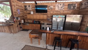 A rustic kitchen with wooden cabinets, a gas stove, a refrigerator, and a bar area with stools. - Avie's Hideaway, Exclusive Private Riverfront @Vacation Frio River Room #1 (Concan)