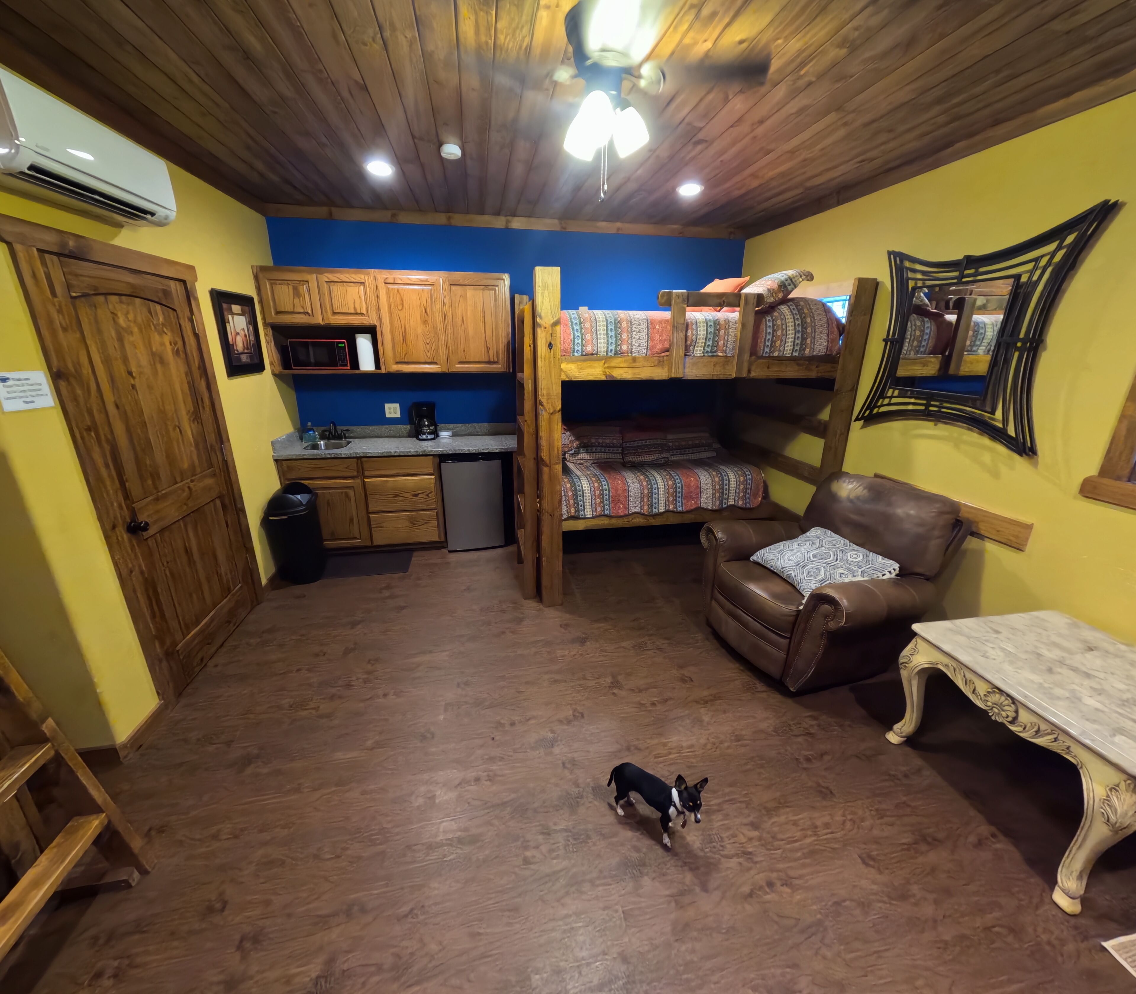 A compact room with wooden flooring, a bunk bed, a sofa, a small table, and a kitchenette area.