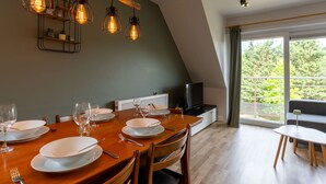 Un comedor con una mesa de madera dispuesta para cuatro, un televisor sobre un mueble y vistas a la vegetación a través de grandes ventanales.