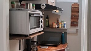 Eine kompakte Küche mit Mikrowelle, Toaster und verschiedenen Küchengeräten. Es gibt ein Fenster mit Jalousien, eine kleine Ablage für Gewürze und einen roten Schrank.