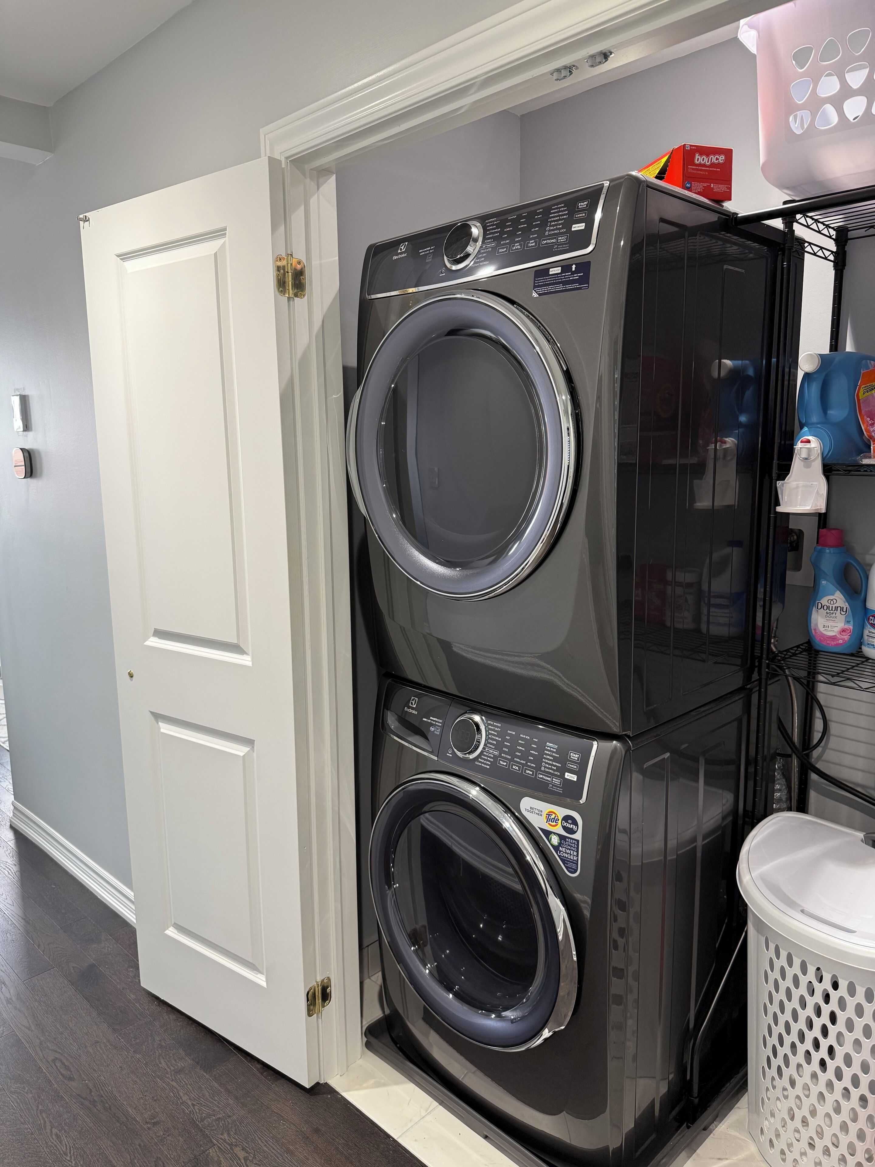 A stacked washer and dryer unit in a laundry room with a white door, a shelf with cleaning supplies, and a trash can.