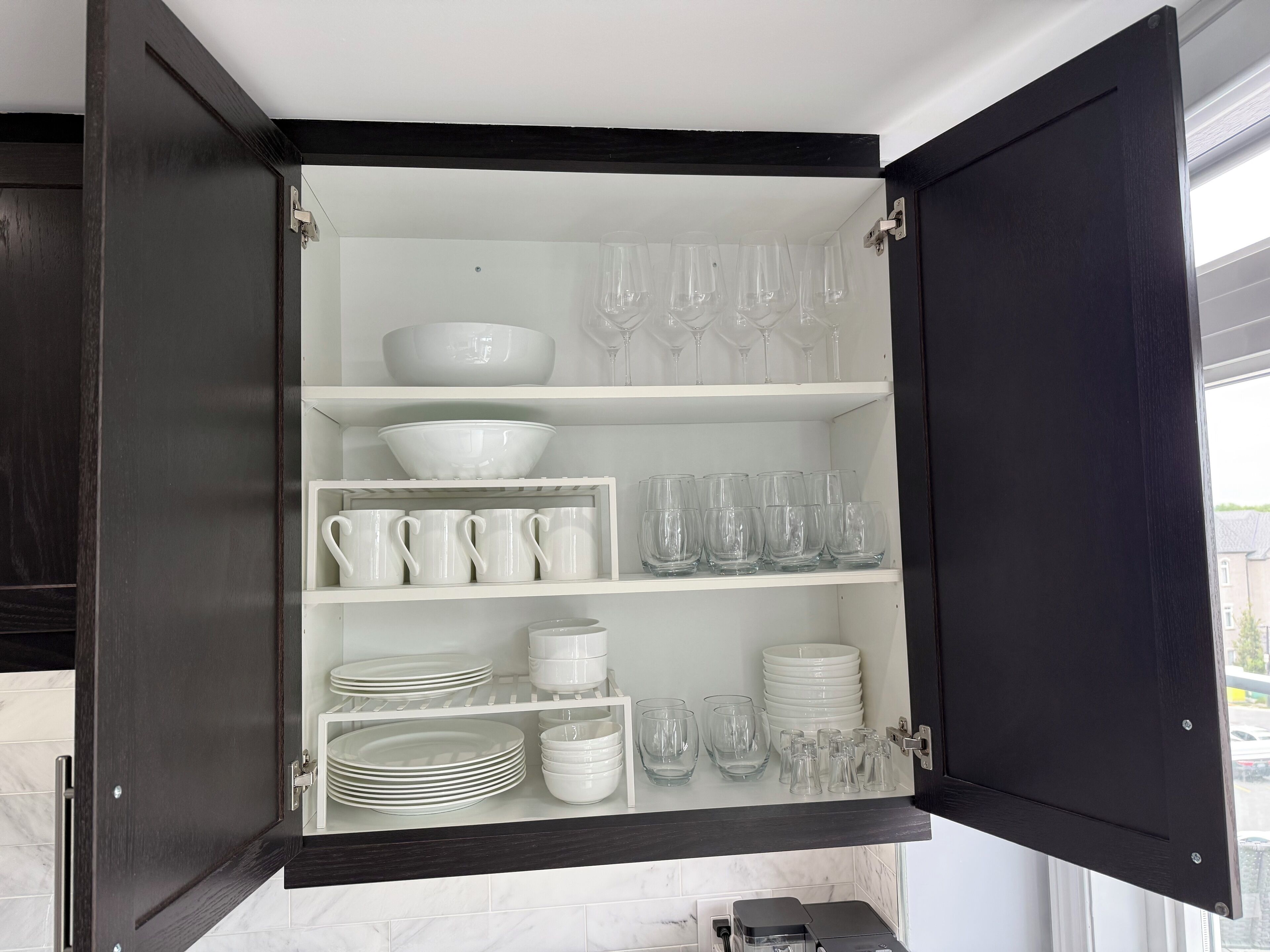 A kitchen cabinet with shelves holding white dishes, glasses, and a coffee maker.