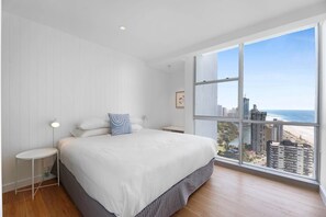 A bedroom with a large bed, a bedside table, a nightstand, a window with a city view, and a ceiling light.