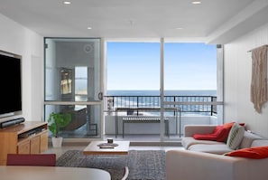 A modern living room with a large TV, a sofa, a coffee table, and a balcony with a view of the ocean.