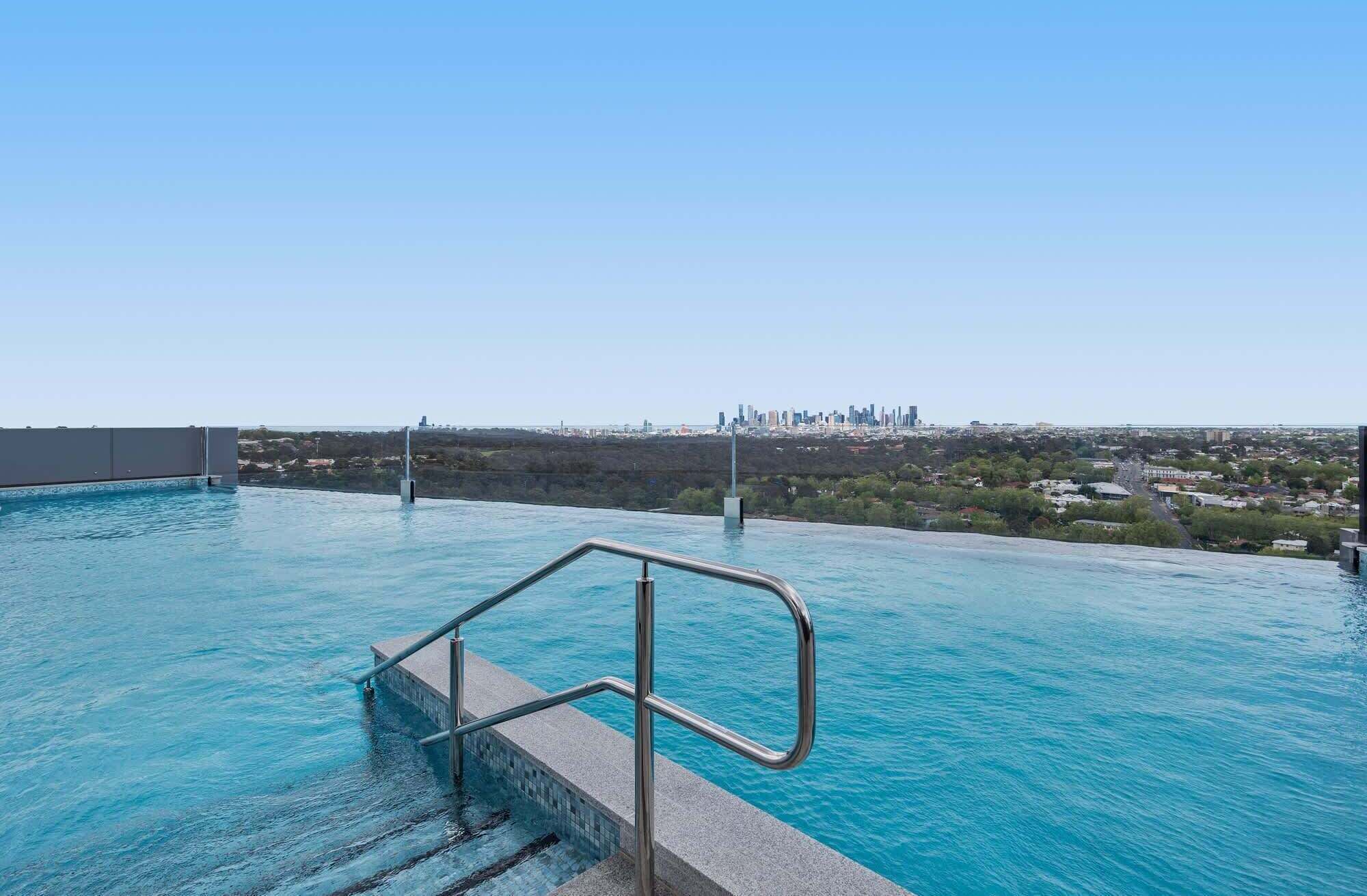 A swimming pool with a metal handrail, clear blue water, and a city skyline in the background.