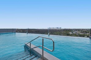 A swimming pool with a metal handrail, clear blue water, and a city skyline in the background.