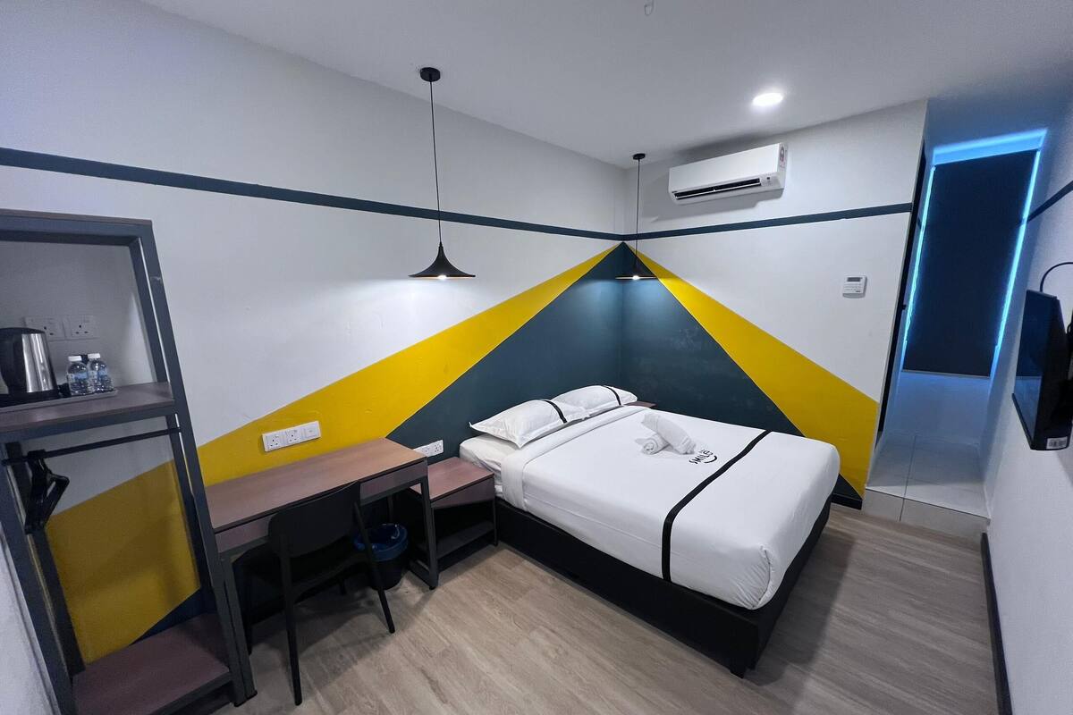 A compact hotel room with a bed, a desk, a chair, and a small table. There is an air conditioning unit on the wall and a door with a blue light.