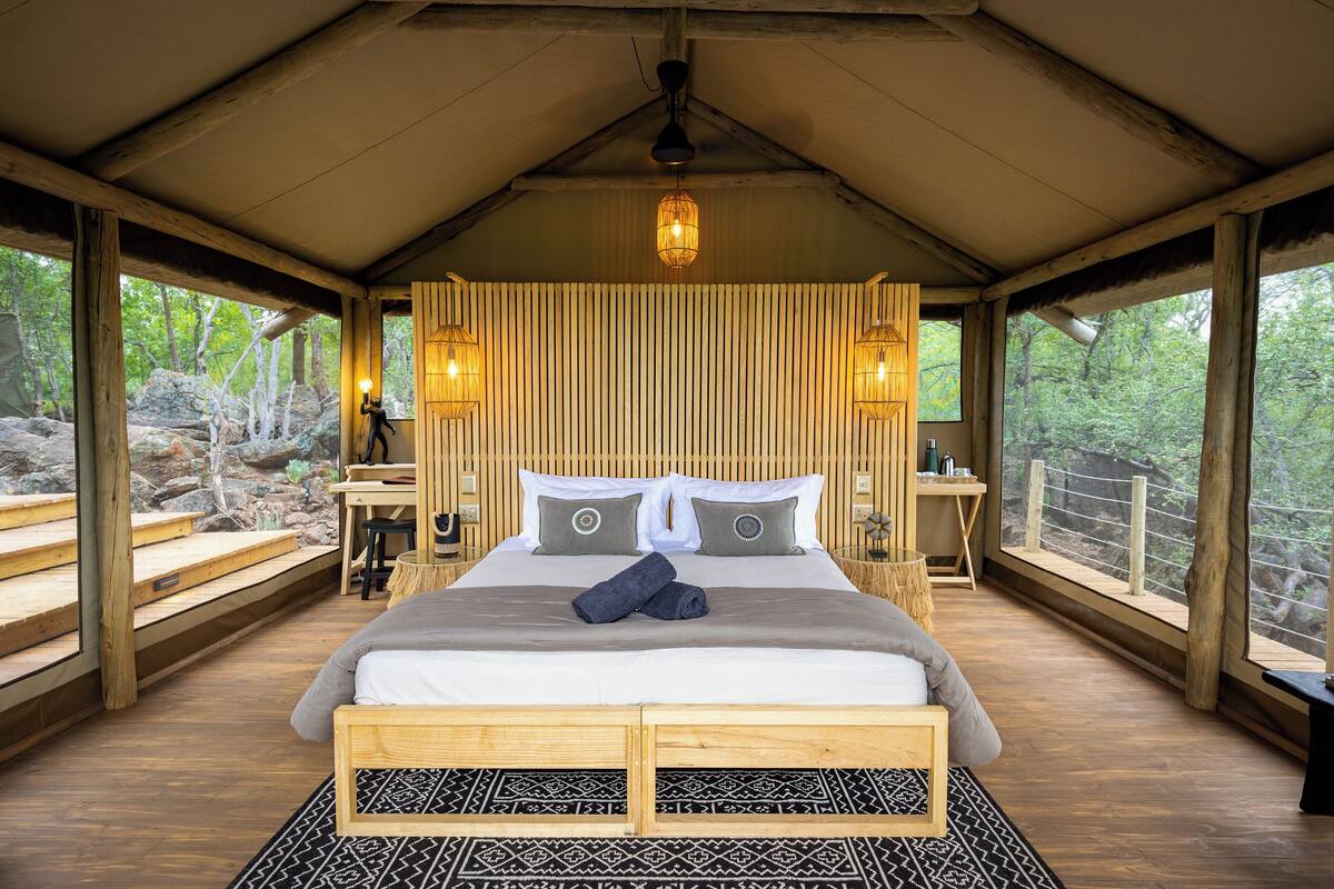 A wooden-framed bedroom with a bed, two bedside tables, a nightstand, and a large window with a view of the outdoors.