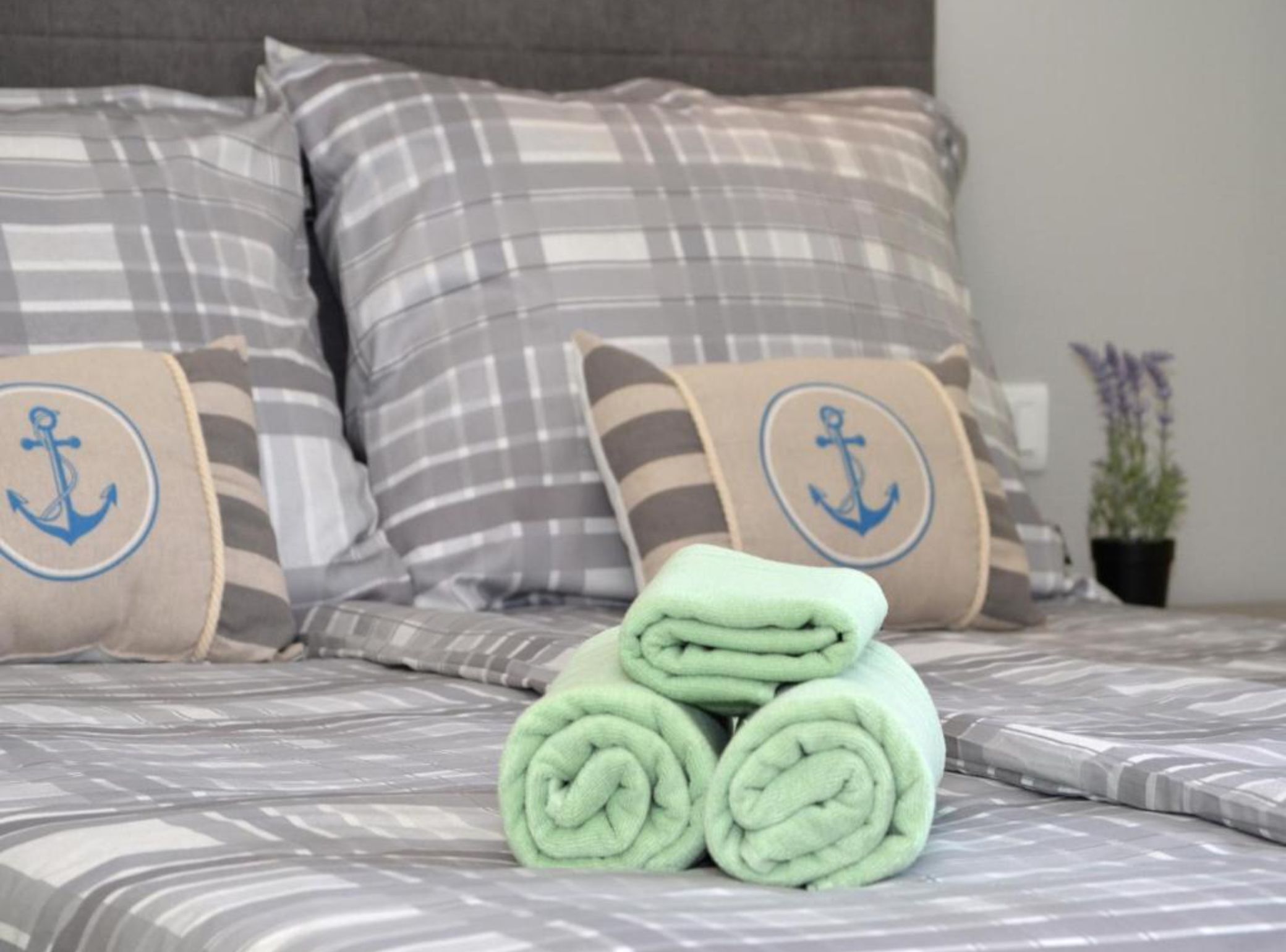A bed with striped bedding and pillows featuring anchor designs, a neatly rolled green towel, and a potted plant on the side.