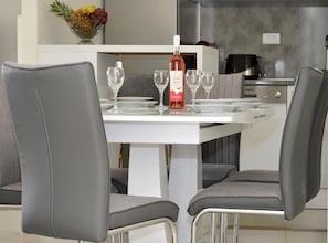 Un comedor moderno con una mesa blanca dispuesta para cuatro personas, que incluye una botella de vino, copas de vino y un cuenco de fruta.