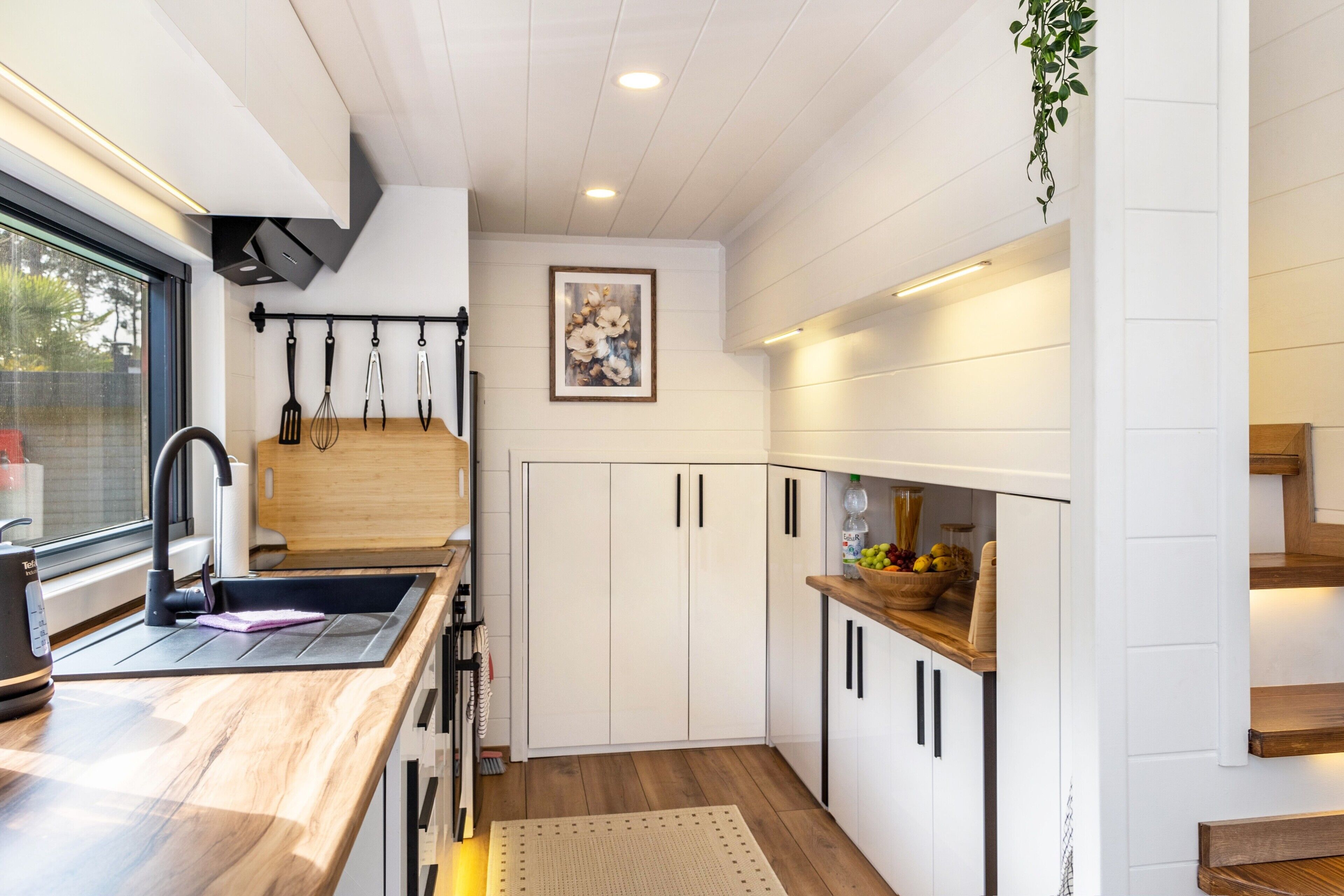 Une cuisine moderne avec un plan de travail en bois, des meubles blancs et un porte-ustensiles suspendu.