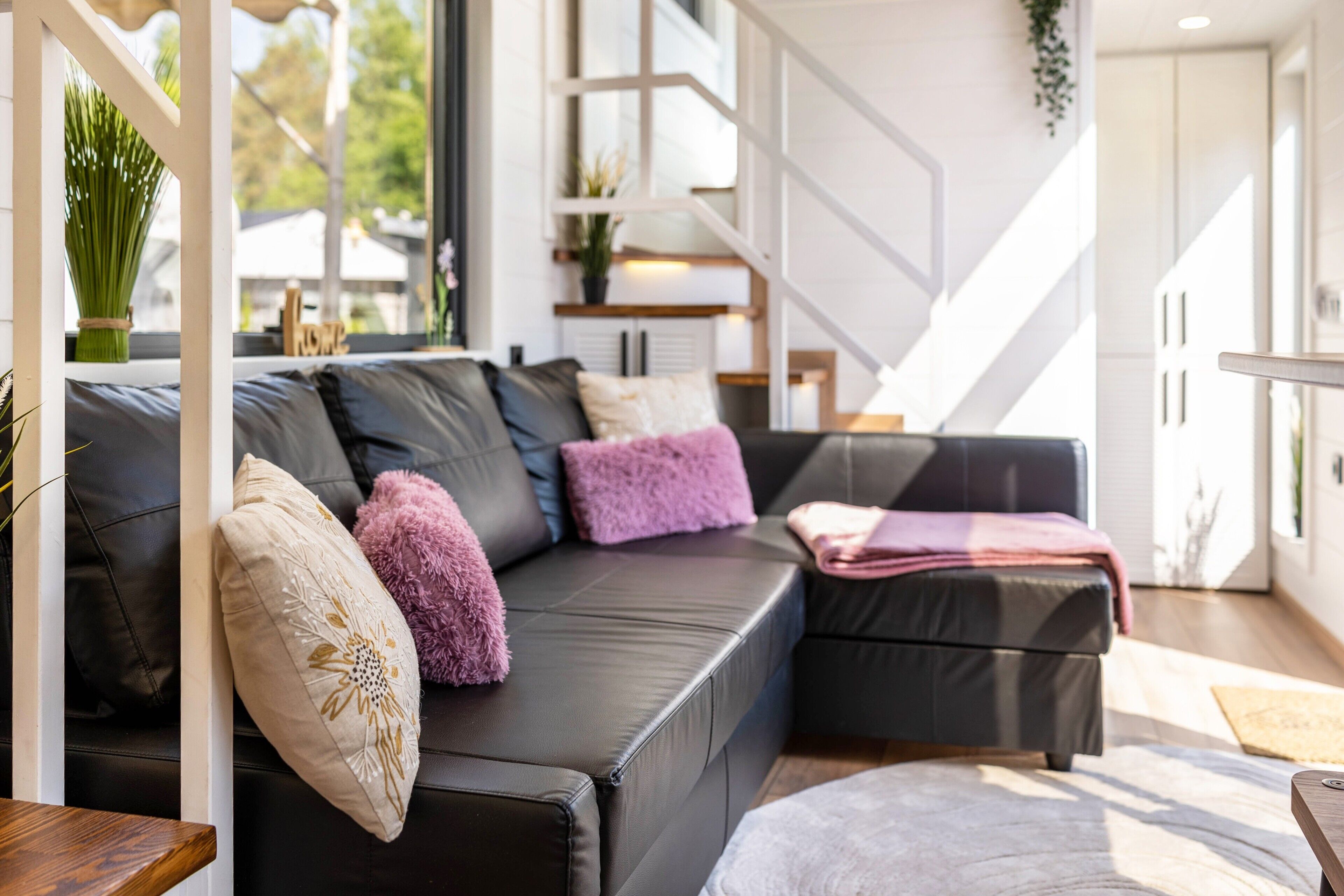 A modern living room with a black leather sofa, decorative pillows, a wooden side table, and a view of a staircase and outdoor greenery.